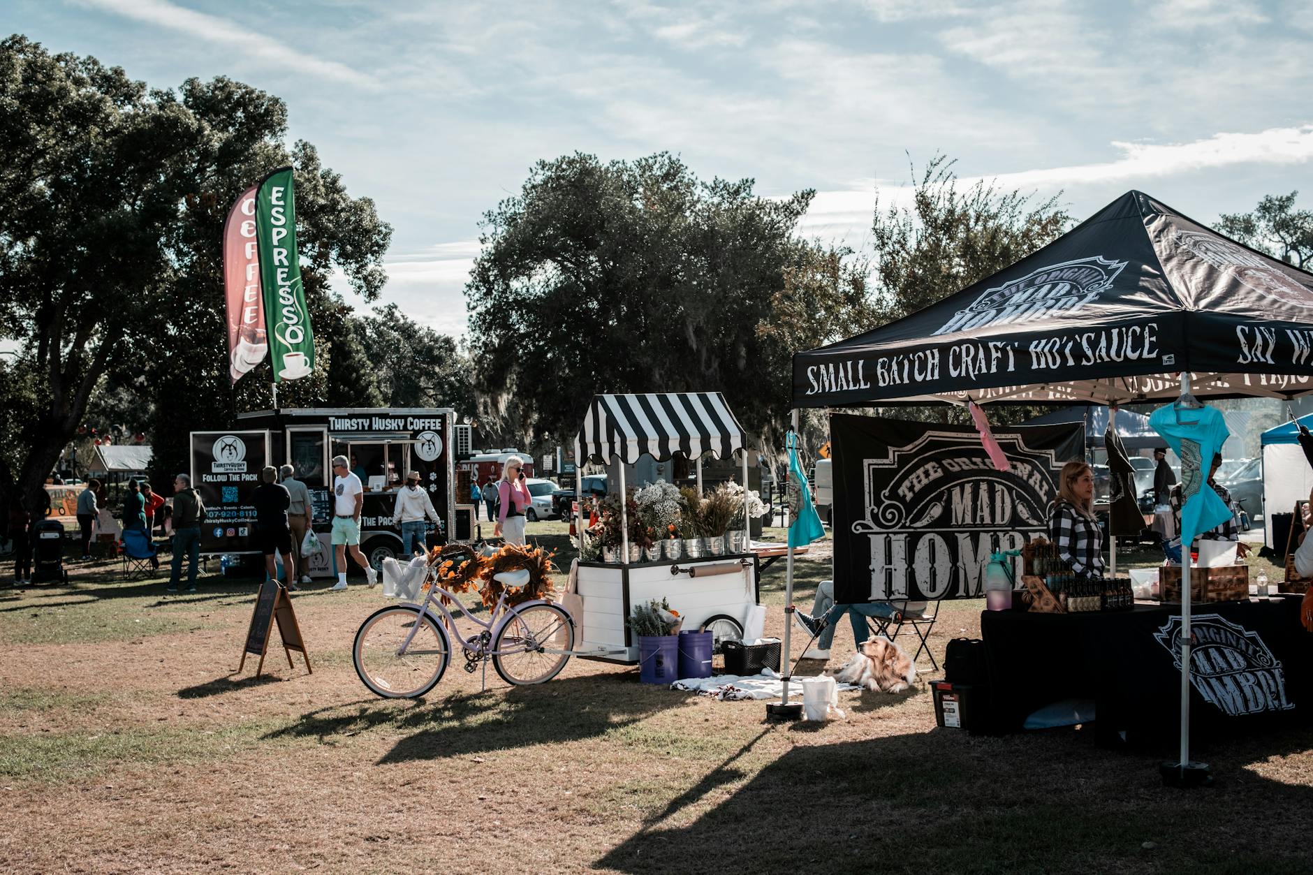 Vendors at a community event with food trucks, tents, and people mingling in a park setting.